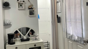 Una cucina compatta con un mobiletto bianco contenente stoviglie, una macchina per il caffè e un bicchiere da vino. Si nota una porta con una tenda a destra.