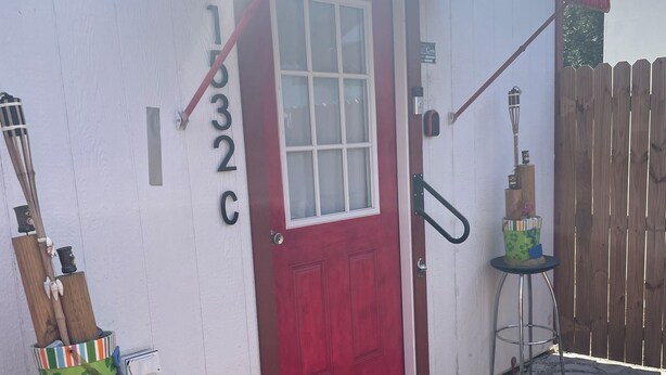 Un edificio con una porta rossa, numero 132C, e un piccolo portico con uno sgabello in metallo e un secchio verde con un bastoncino di legno.