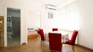 A room with a wooden floor, a round table with floral tablecloth, red chairs, a bed with a pink blanket, a shower area, and a window with curtains. - Apartment 1362/116992 (Istria - Peroj), 500m from the beach (Peroj)