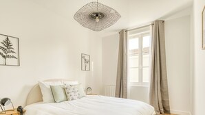 A bedroom with a bed, a bedside table, a lamp, a wall-mounted light fixture, and a window with curtains.