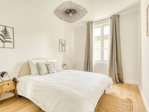 Un dormitorio con una cama, una mesita de noche, una lámpara, un aplique de pared y una ventana con cortinas.