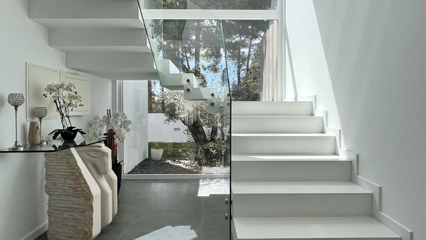 A modern interior with a glass staircase leading to a balcony, a minimalist living area with a sofa and decorative items, and a view of trees outside. - Villa do Mar - Close to the beach (Aroeira)
