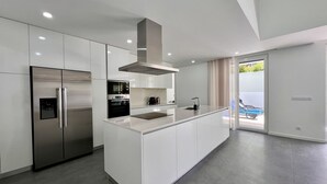 A modern kitchen with white cabinetry, a stainless steel refrigerator, an oven, and a built-in island with a sink. - Villa do Mar - Close to the beach (Aroeira)
