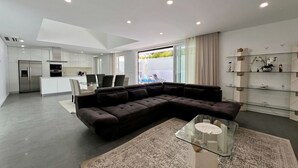 A modern living room with a dark brown sectional sofa, a glass coffee table, and a patterned rug. - Villa do Mar - Close to the beach (Aroeira)