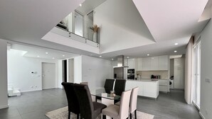 A spacious, modern kitchen with a central island, a dining area with a glass table and chairs, and a high ceiling with skylights. - Villa do Mar - Close to the beach (Aroeira)