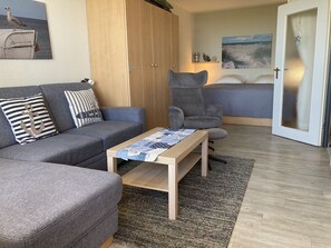 Un salon moderne avec un canapé gris, un fauteuil assorti, une table basse en bois et une grande fenêtre donnant sur la mer.