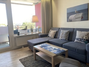 Un salon moderne avec un canapé gris, une table basse en bois et une vue sur la mer par une grande fenêtre.