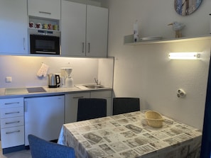 Une cuisine compacte équipée d’un four encastré, d’une machine à café et d’un évier. On y trouve une petite table à manger, recouverte d’une nappe à motifs de journaux, ainsi qu’un panier.