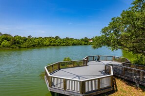 A wooden deck with a railing overlooking a calm lake, surrounded by lush greenery.