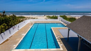 Une piscine rectangulaire à l’eau d’un bleu cristallin, entourée d’une clôture blanche, avec une plage et une vue sur l’océan en arrière-plan.