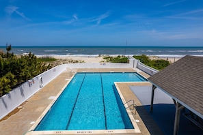 A rectangular pool with clear blue water, surrounded by a white fence, with a beach and ocean view in the background.