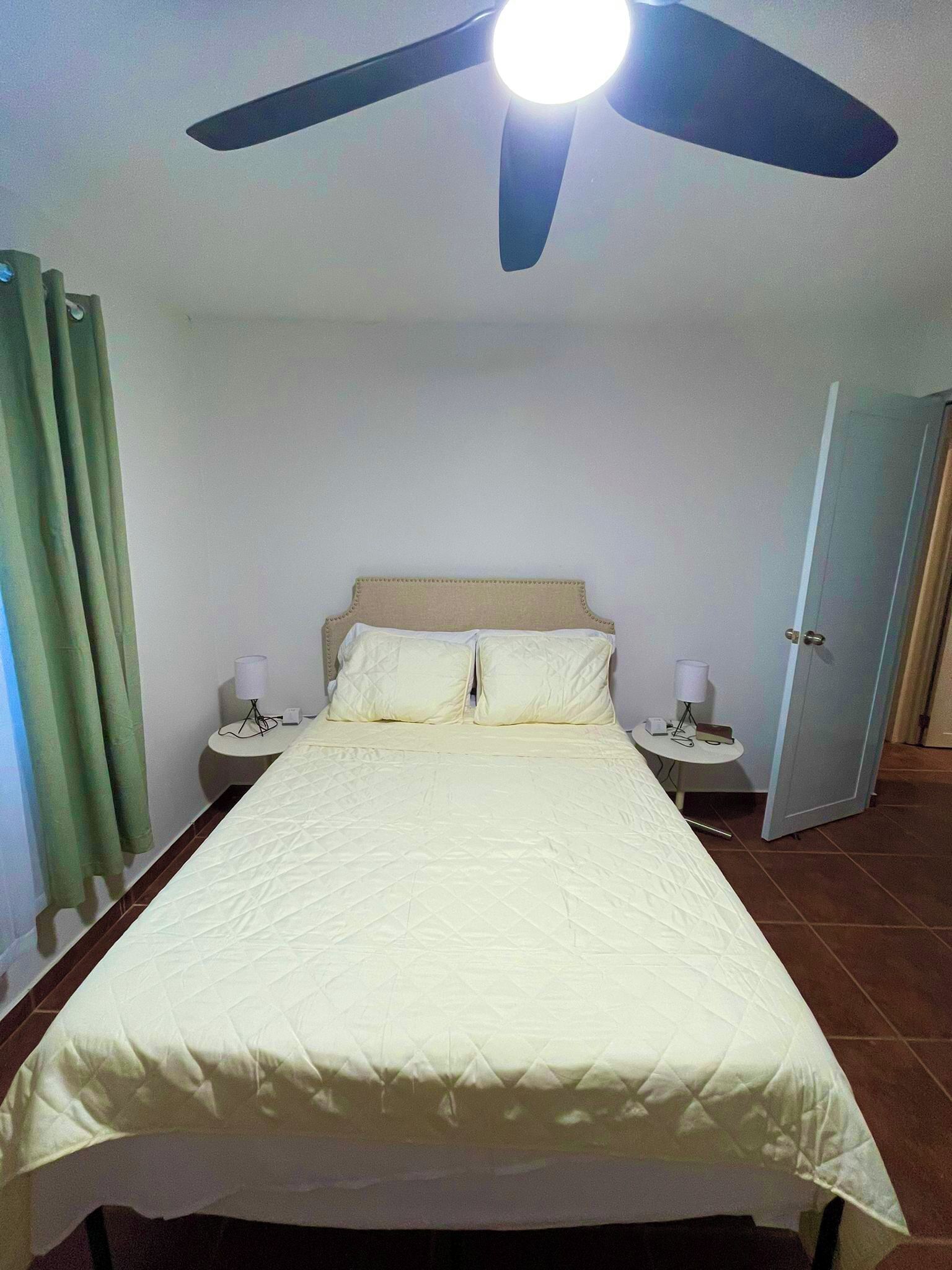 Una habitación de hotel con cama, ventilador de techo, mesita de noche con lámpara y puerta.