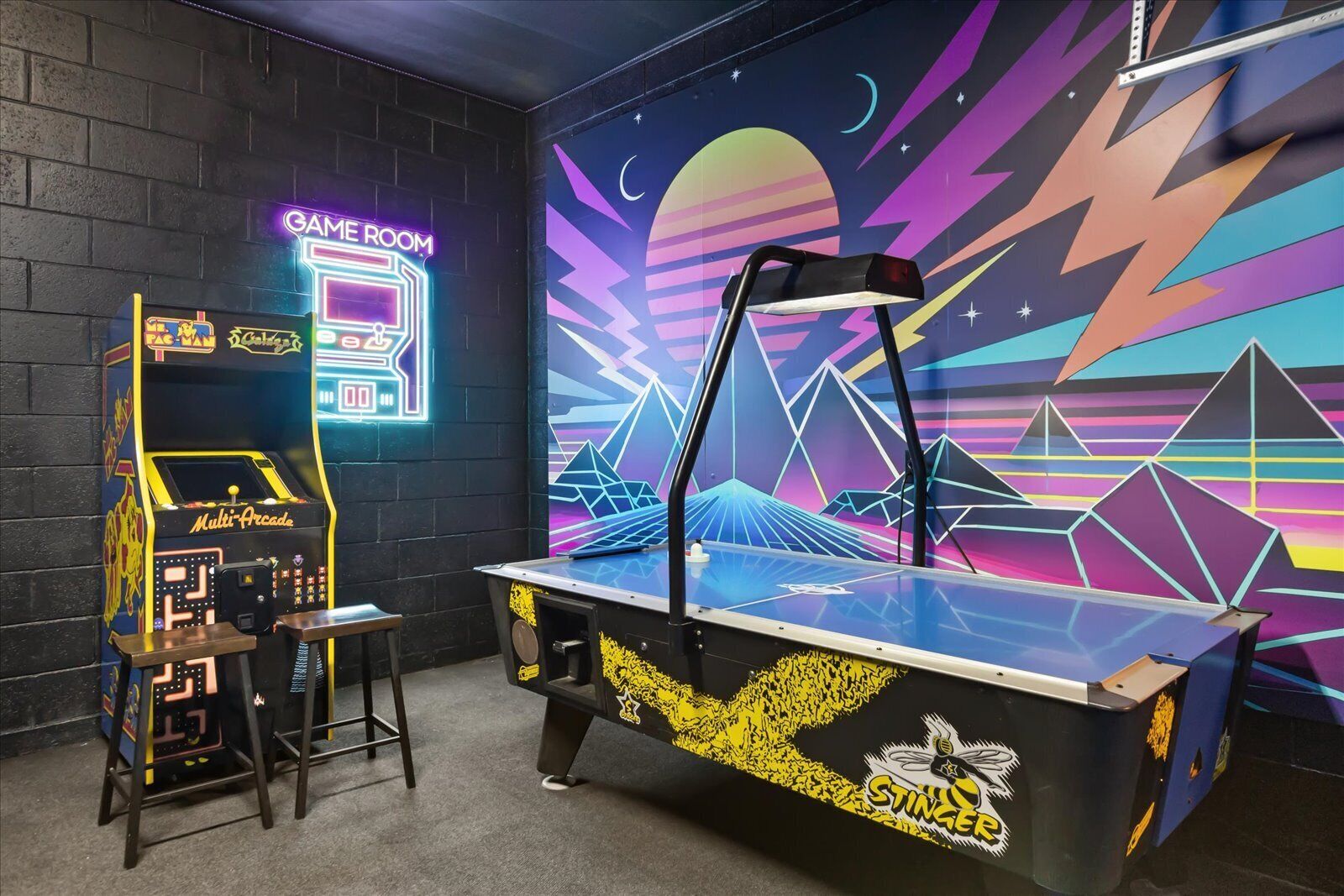 A neon-lit arcade room with a multi-arcade game machine and a table tennis table against a vibrant geometric wall.
