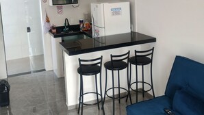 A compact kitchen with a black countertop, three black bar stools, a white refrigerator, and a microwave above the counter. - Pousada da Família com Apartamentos Mobiliados e Completos! (Santa Lidia Penha)