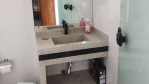 A bathroom with a granite sink, a mirror, a glass shower enclosure, and a trash bin. - Pousada da Família com Apartamentos Mobiliados e Completos! (Santa Lidia Penha)
