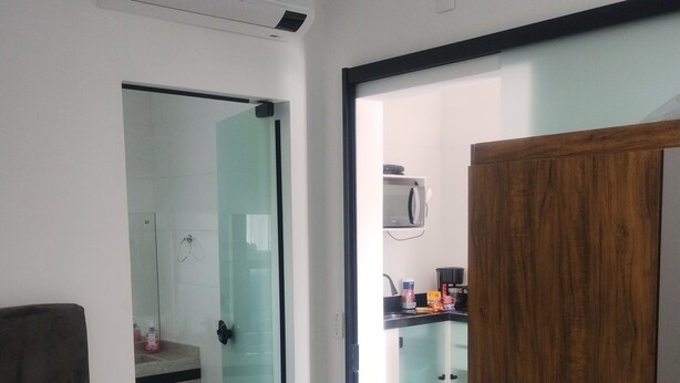 A modern hotel room with a bed, a wooden wardrobe, a glass door leading to a bathroom, and a kitchenette area. - Pousada da Família com Apartamentos Mobiliados e Completos! (Santa Lidia Penha)