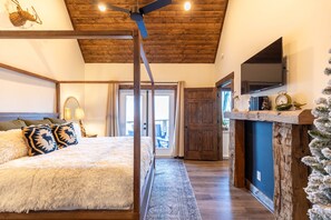 Une chambre avec un grand lit, un plafond en bois, un téléviseur fixé au mur et une cheminée rustique.