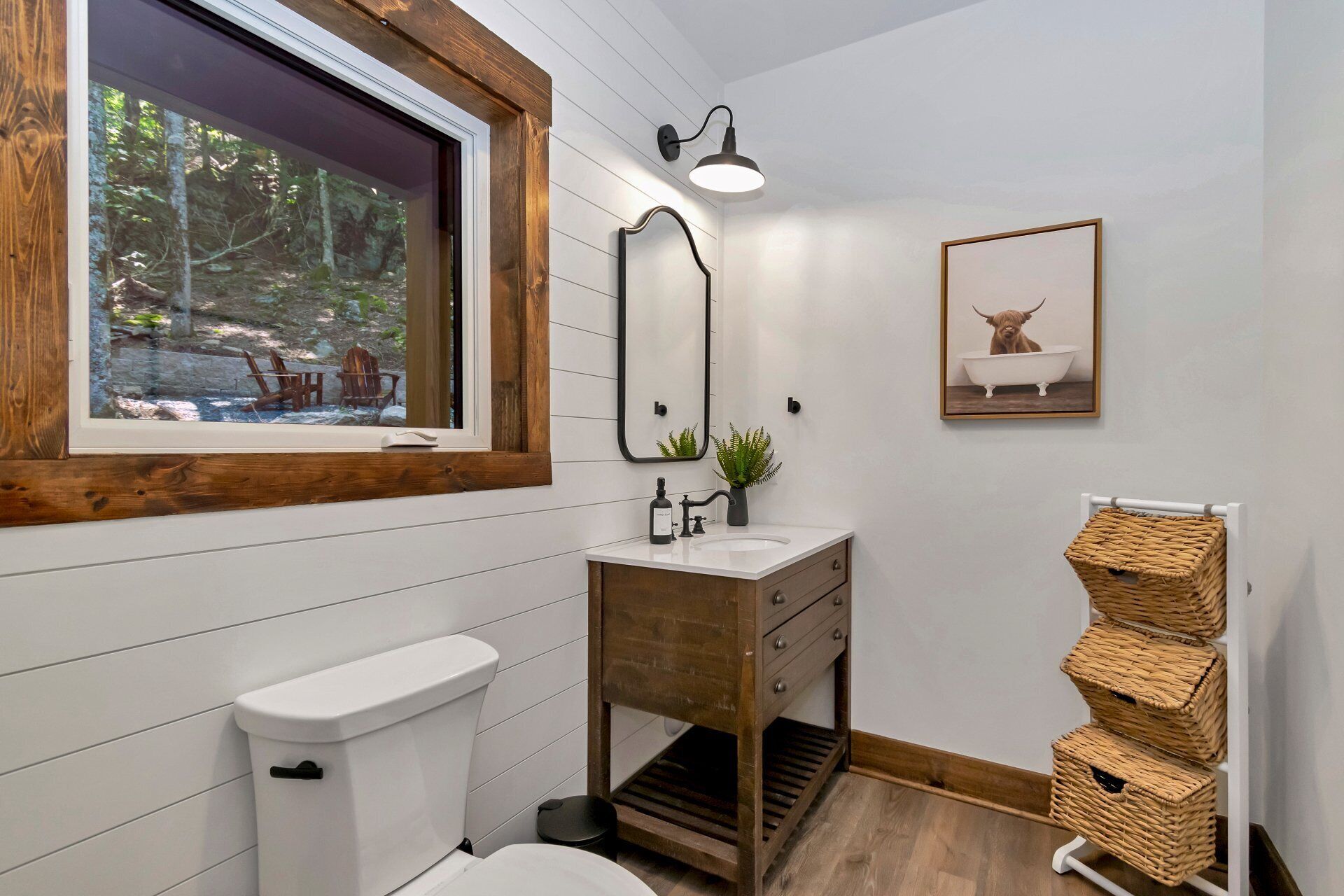 Ein Badezimmer mit Holzwaschtisch, Toilette in Weiß, Fenster mit Blick auf einen Wald, einer Wandleuchte und einem Bilderrahmen mit dem Motiv eines Bullen in einer Badewanne.