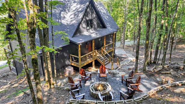 A cabin with a deck, surrounded by trees, and a fire pit with chairs.