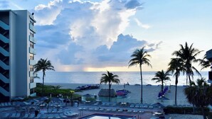 A resort pool with a view of the ocean and beach, surrounded by palm trees and lounge chairs.