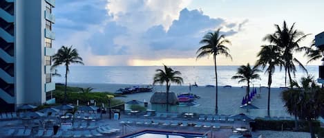 A resort pool with a view of the ocean and beach, surrounded by palm trees and lounge chairs.