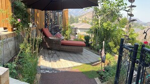 Un fauteuil de jardin en osier avec un coussin rouge, placé sous un grand parasol, un jardin avec différentes plantes et une vue sur un paysage montagneux.