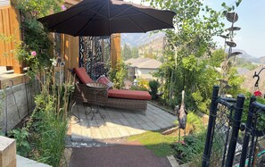 A wicker lounge chair with a red cushion under a large umbrella, a garden with various plants, and a view of a mountainous landscape. - Shannon Ridge Mountain View Home (West Kelowna)