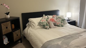 Un dormitorio con cama, mesita de noche, lámpara, almohada decorativa, un florero con flores y un mueble de almacenamiento con cestas.