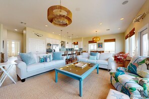 A spacious living room with a large coffee table, white sofas, and a colorful armchair. A kitchen is visible in the background with modern appliances.