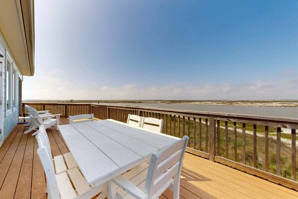 A wooden deck with a white table and chairs, overlooking a beach and ocean.