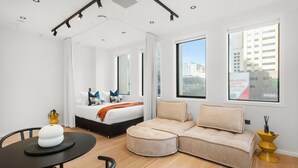 A modern hotel room with a large bed, a beige sofa, a dining table, and a city view through the windows. - Victoria Street Apartments (Auckland)