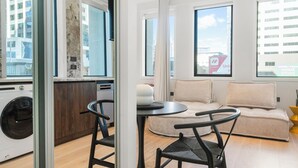 A modern living room with a sofa, a dining table, and chairs. There is a kitchen area with a washing machine visible through a glass door. - Victoria Street Apartments (Auckland)