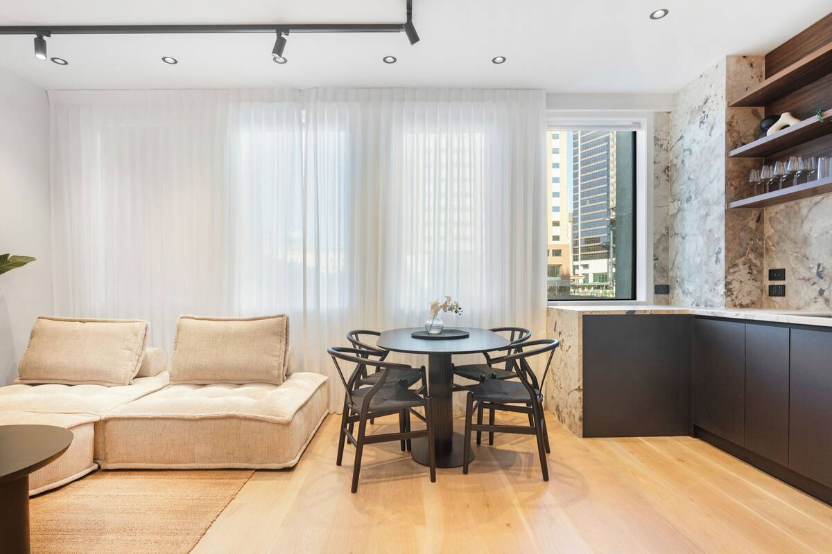 A modern living room with a round dining table, dark wooden chairs, a beige sofa, and a large window with sheer curtains.