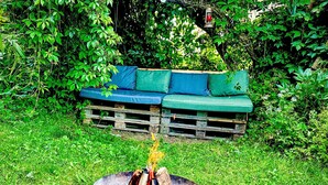 Un brasero con leña en un contenedor metálico circular, rodeado de césped. En el fondo, un conjunto de sofás verdes sobre un palet de madera, cubierto parcialmente por un lona verde.