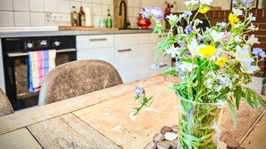Una cocina con armarios blancos, salpicadero de azulejos, una mesa de madera con un florero de vidrio y una pared de ladrillo al fondo.