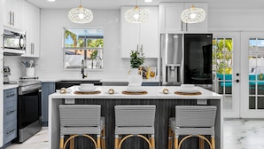 A modern kitchen with white cabinetry, a central island with bar stools, stainless steel appliances, and decorative hanging lights.