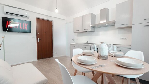 A modern kitchen with white cabinets, a wooden dining table set for four, and a flat-screen TV mounted above the door. - [Torino Centro] Odissea House (Torino)