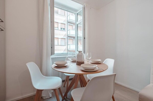 Un coin repas avec une table ronde dressée pour quatre personnes, des chaises blanches et une vue sur un bâtiment par la fenêtre.