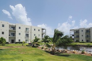 Un edificio residencial de varios pisos con balcones, un césped y una pequeña laguna.