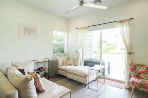 A well-lit living room with a sofa, a coffee table, a side table, and a chair. There is a window with curtains, a ceiling fan, and a door leading outside. - Modern 2 bed with pool and gym (West Bay)
