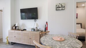 Uma área de refeições com mesa redonda, cadeiras, televisão fixada na parede e um pequeno armário com objetos decorativos.