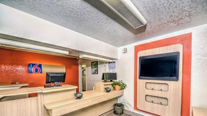 A hotel reception area with a wooden reception desk, a digital screen, a sign indicating room number 6, and a wall-mounted TV. - Motel 6 – Colorado Springs, CO – South/Metro Area (Colorado Springs)