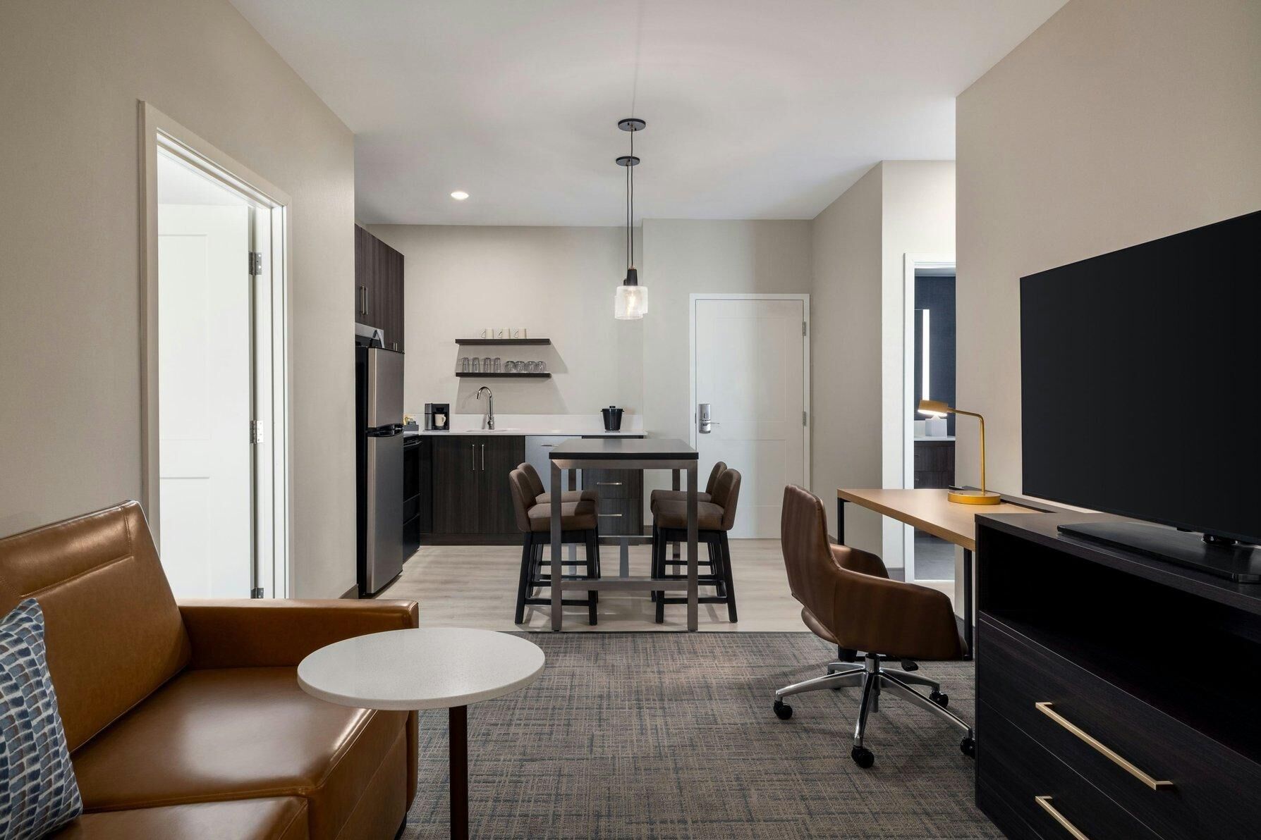A compact hotel room with a kitchenette, a dining area, a TV, and a seating area with a sofa and chairs.