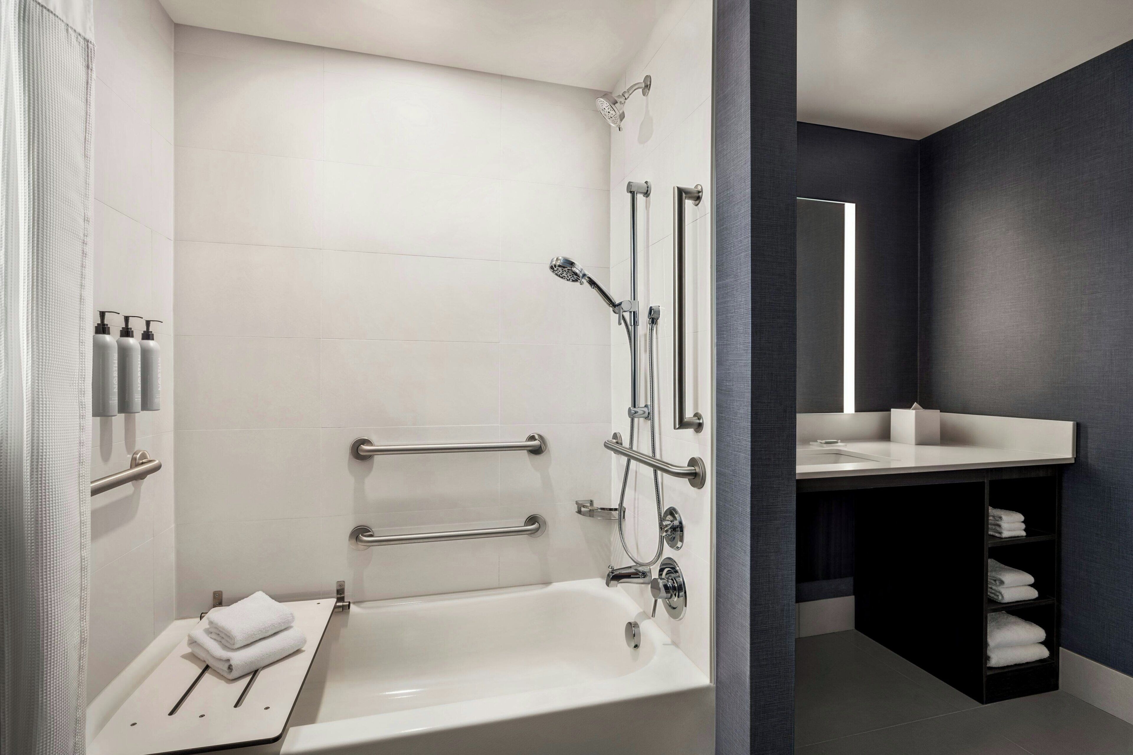 A modern bathroom with a bathtub, grab bars, a showerhead, and a vanity with a mirror.