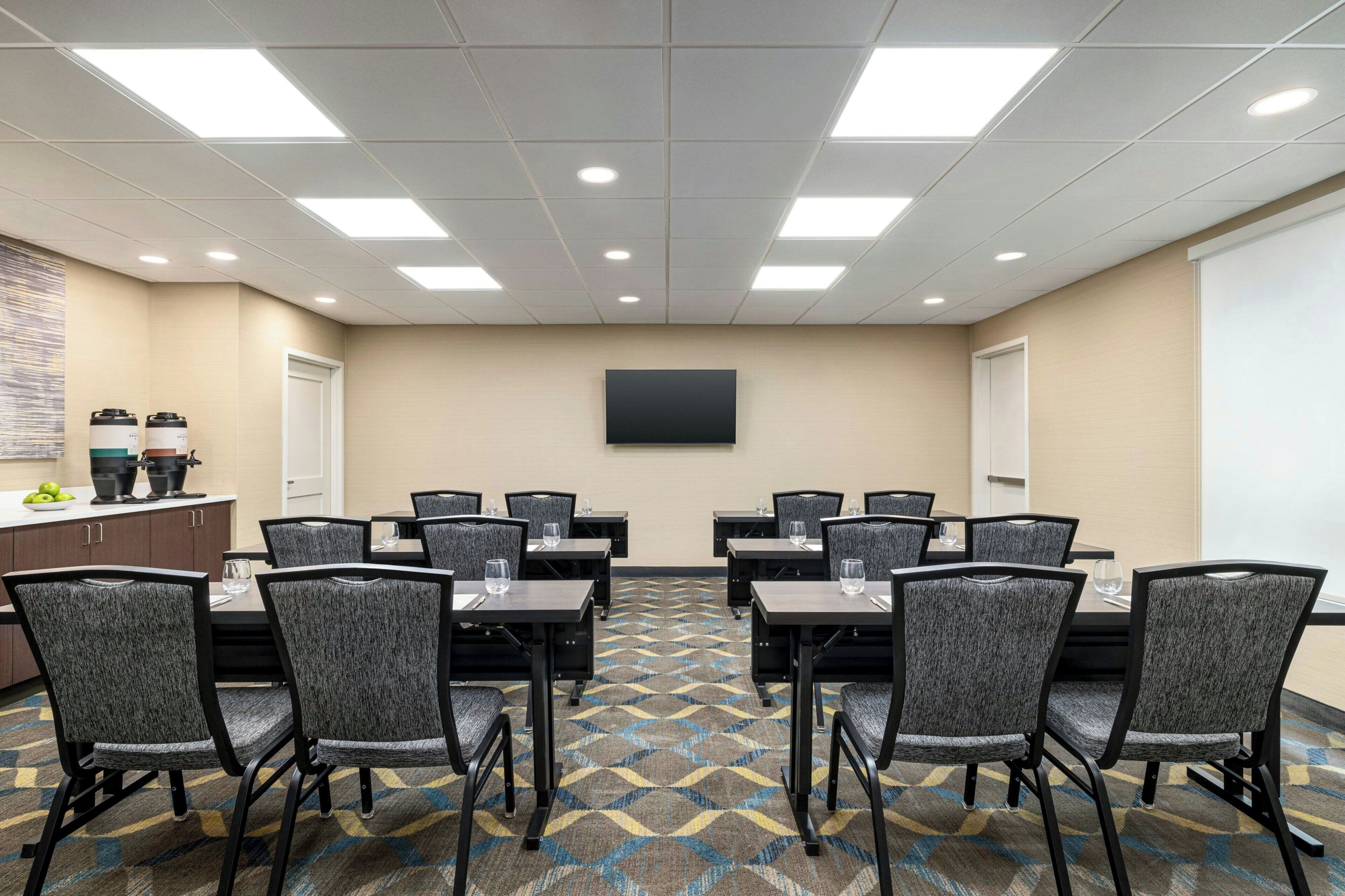 A conference room with a large screen, multiple tables with chairs, and a patterned carpet.