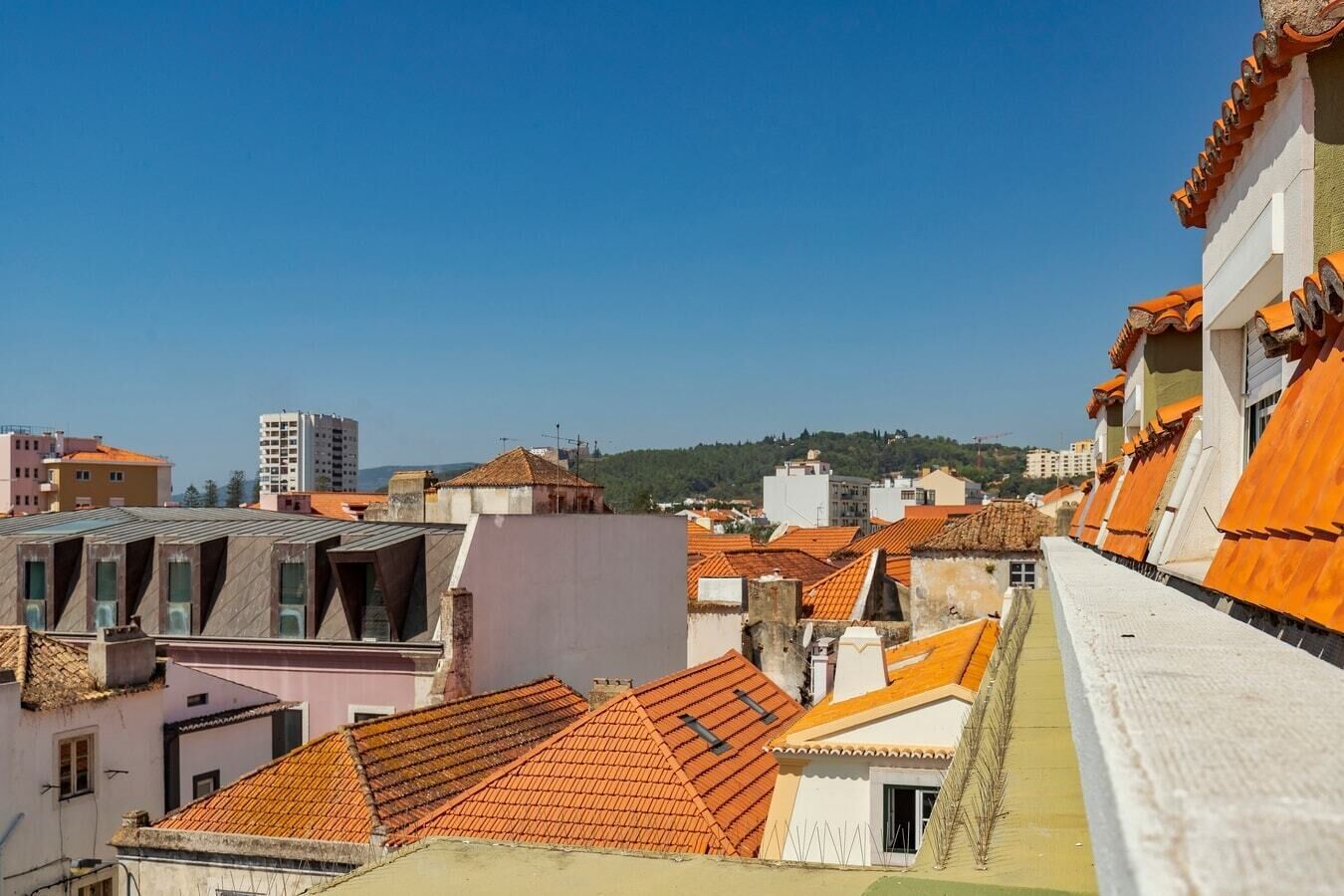 A cityscape with terracotta-tiled rooftops, a mix of residential and commercial buildings, and a clear blue sky.