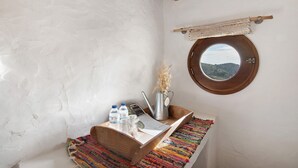 Una habitación rústica con un banco de madera, una mesita con un florero y botellas, y un ventanal con vistas.