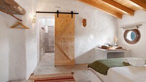Un dormitorio con puerta de madera, una cama con manta verde, una mesita con artículos de aseo y un espejo redondo en la pared.