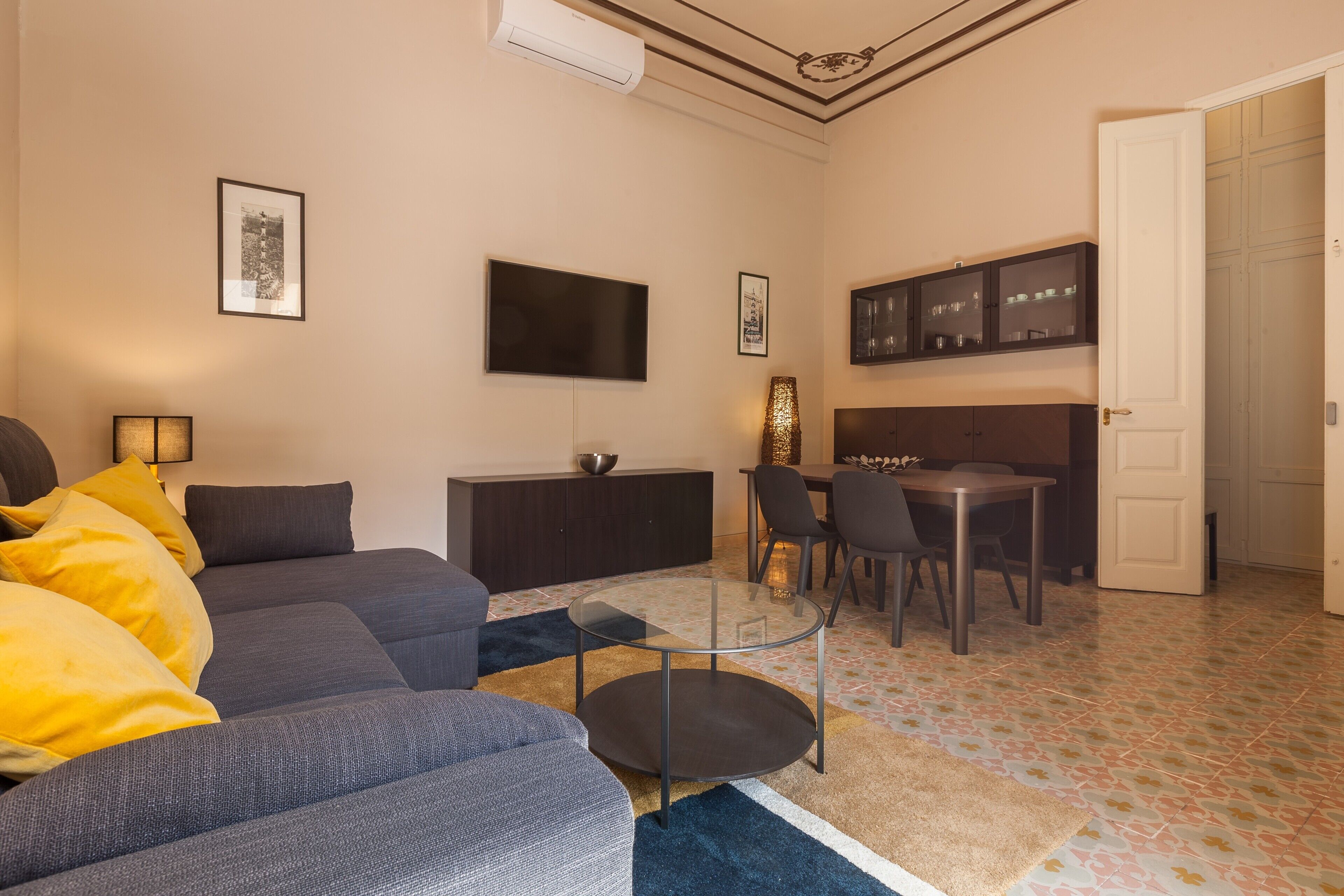 Een woonkamer met een bank, een glazen salontafel, een televisie, een eethoek met stoelen, en een keuken met kasten.