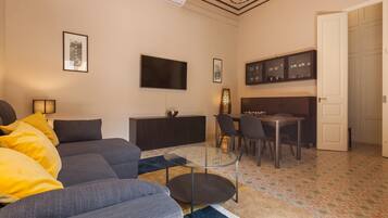 Een woonkamer met een bank, een glazen salontafel, een televisie, een eethoek met stoelen, en een keuken met kasten.